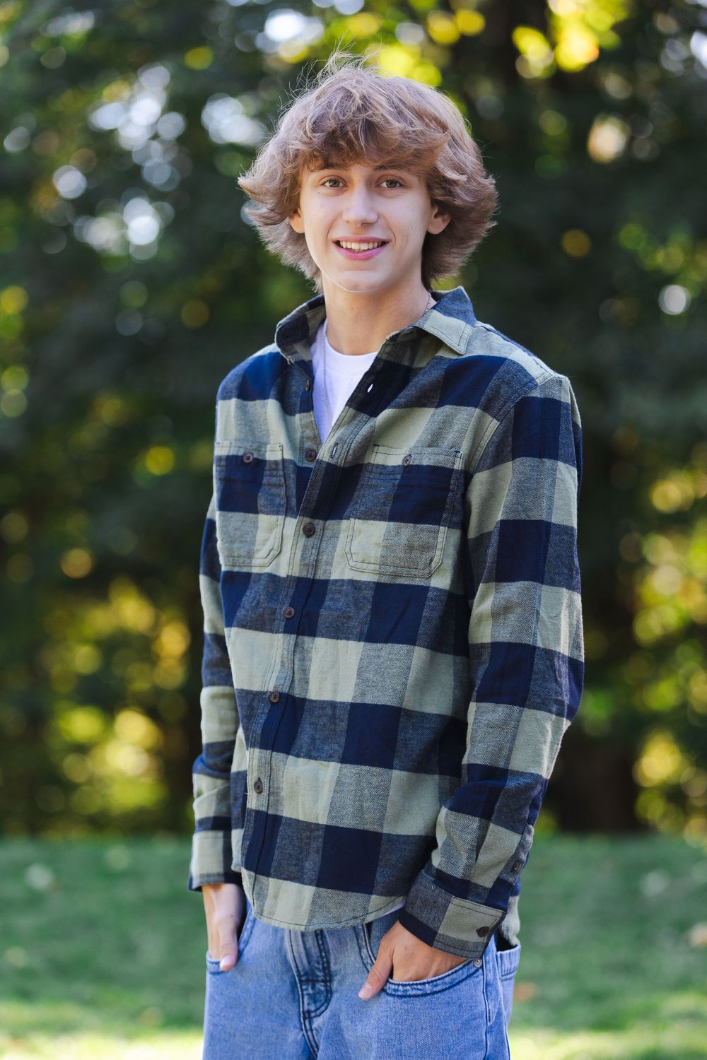 Teenager in plaid flannel shirt posing outdoors with autumn foliage in background.