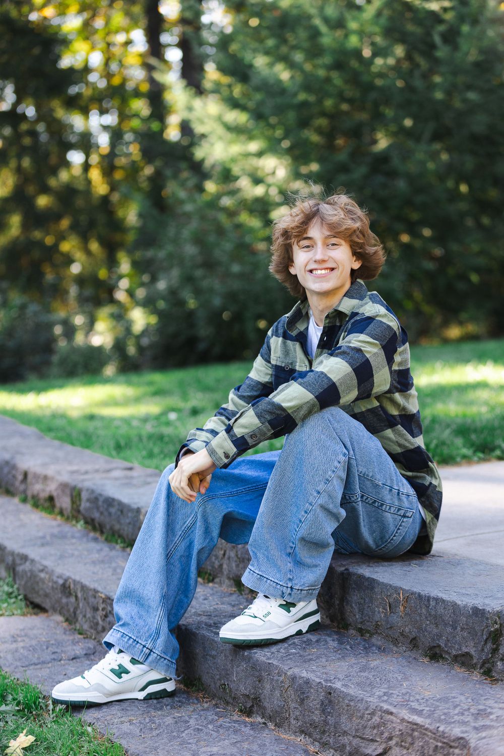 Person in plaid shirt and jeans sitting casually on concrete steps outdoors.