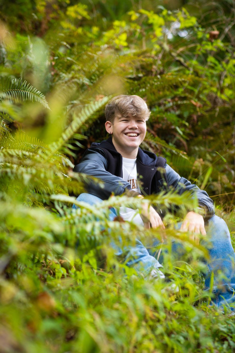 Person in blue jacket sits among lush green ferns and foliage.