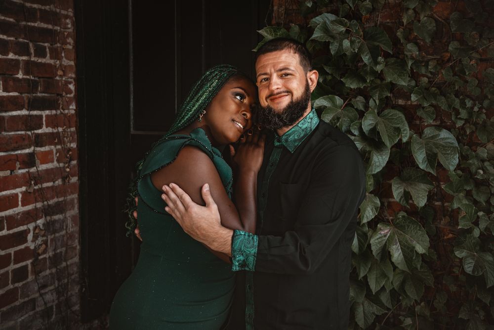 An intimate couple shares a close embrace against a brick wall covered in green vines.