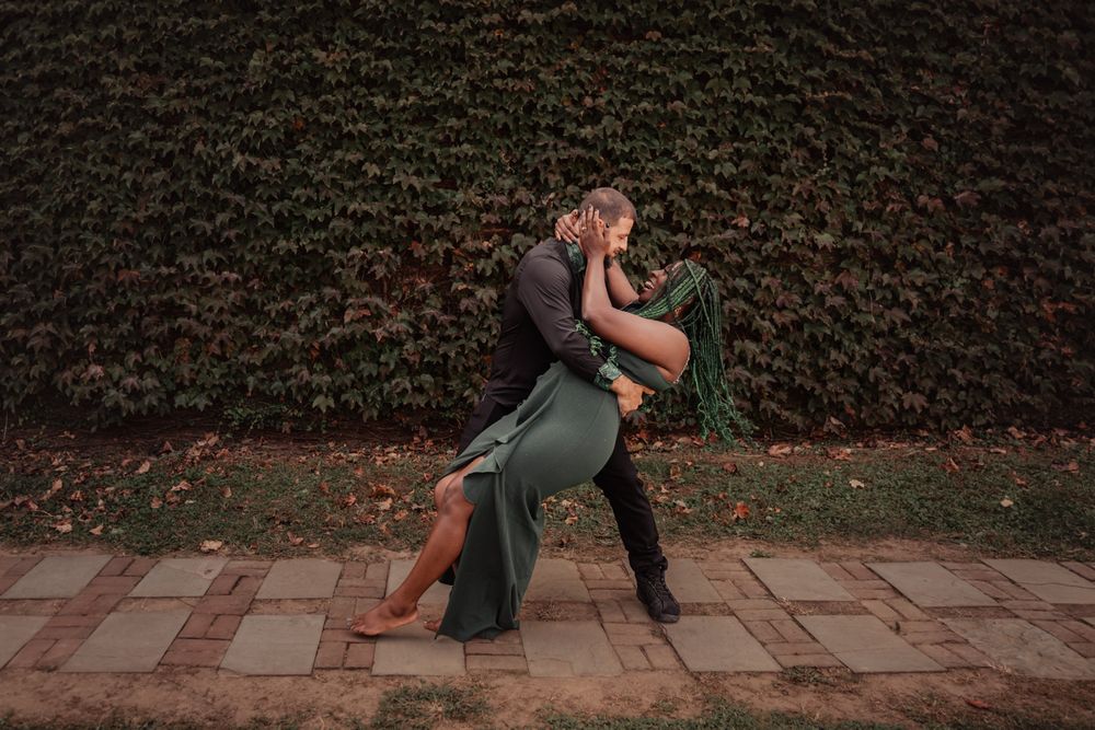 A romantic couple embraces and dances together against a brick wall with climbing vines in an outdoor setting.
