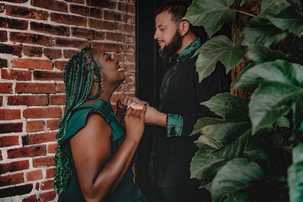 A couple shares a tender moment against a rustic brick wall with green plants.