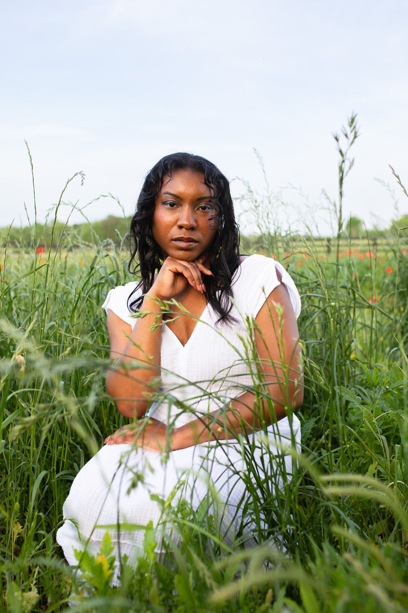 A portrait in a serene meadow setting with tall grass and wildflowers features a woman in a white dress against a soft sky.