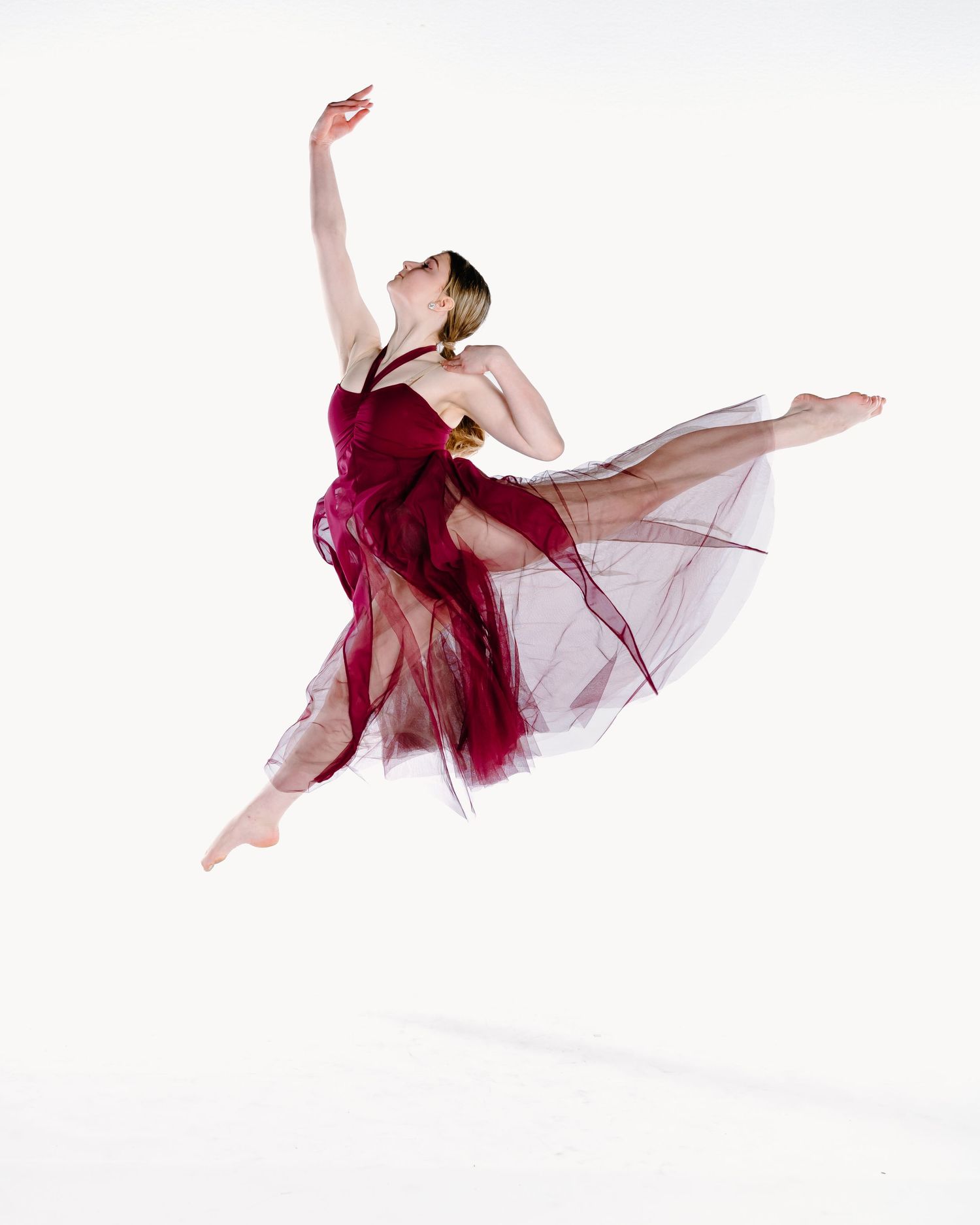 Dancer in flowing burgundy dress and sheer fabric performs graceful leap against white background.
