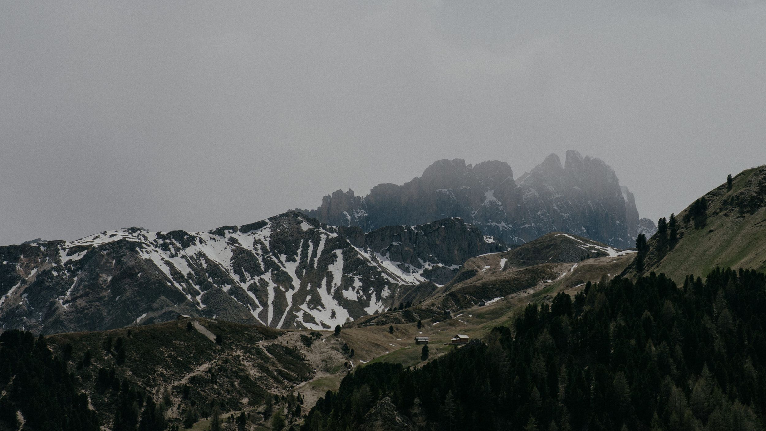 Dramatic mountain landscape with snow-capped peaks and green slopes under a moody overcast sky in the Dolomites.
