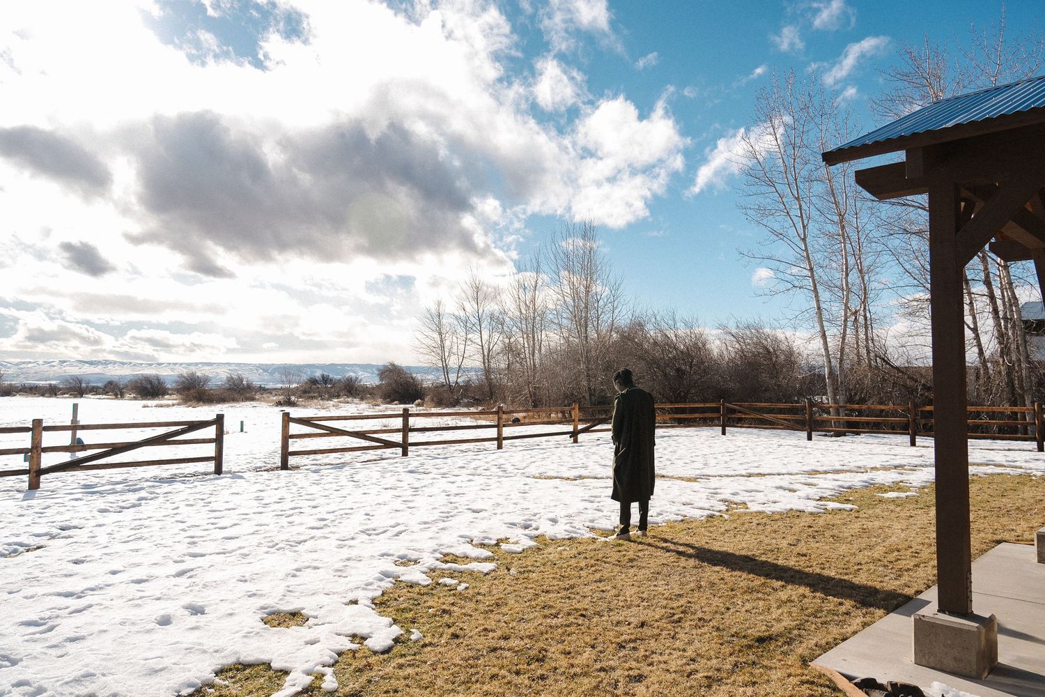 A snowy winter landscape with wooden fences and bare trees under cloudy skies and patches of exposed ground.