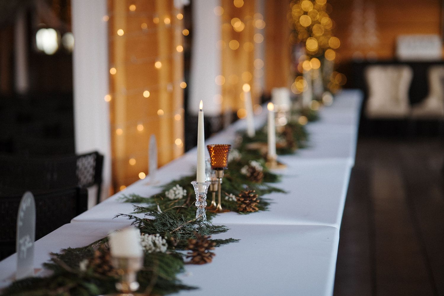 Elegant table setting with white linens, evergreen garland, and glowing string lights creating a romantic ambiance.