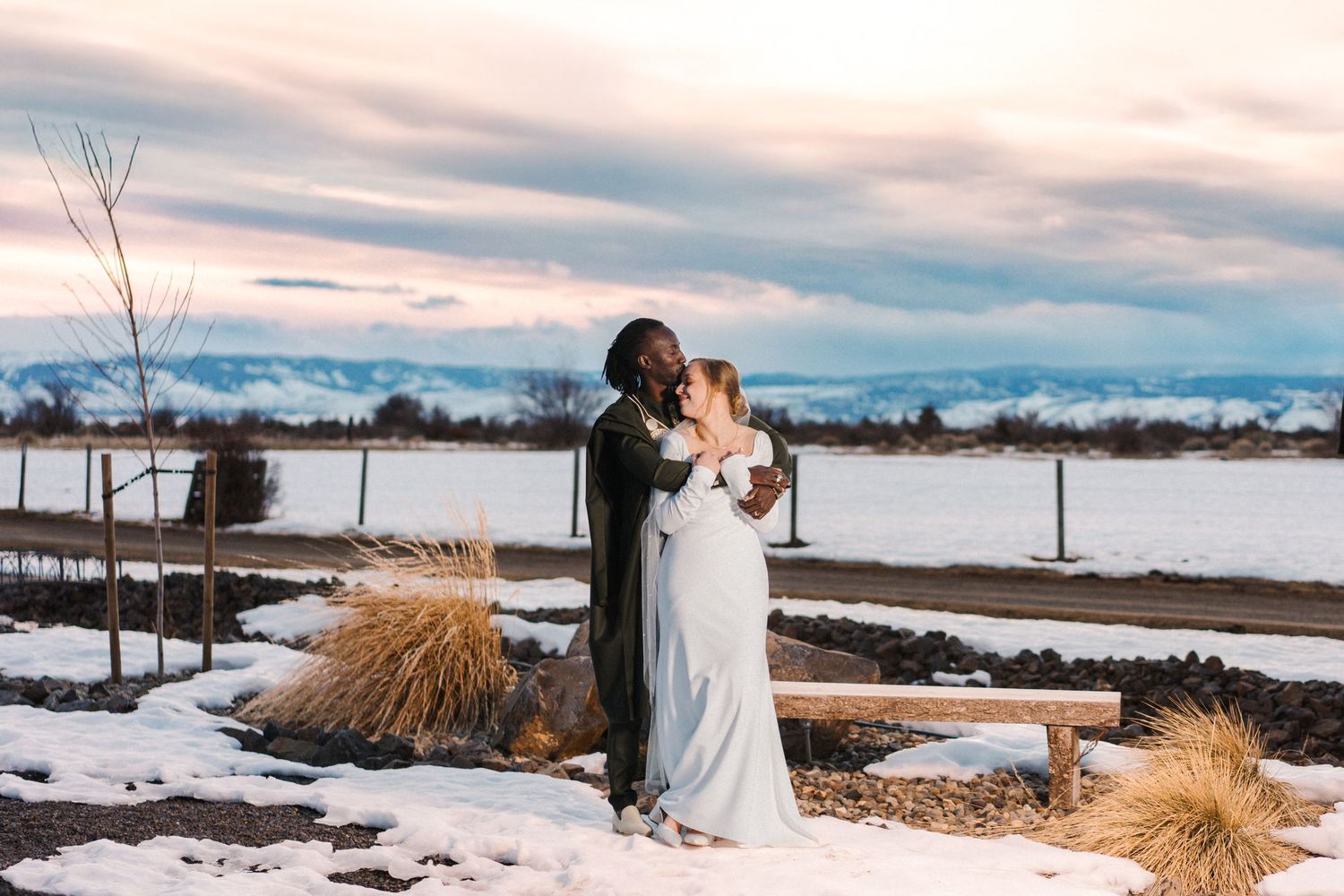 A couple shares a romantic winter wedding moment on snowy terrain with dried grasses and a cloudy sunset sky.