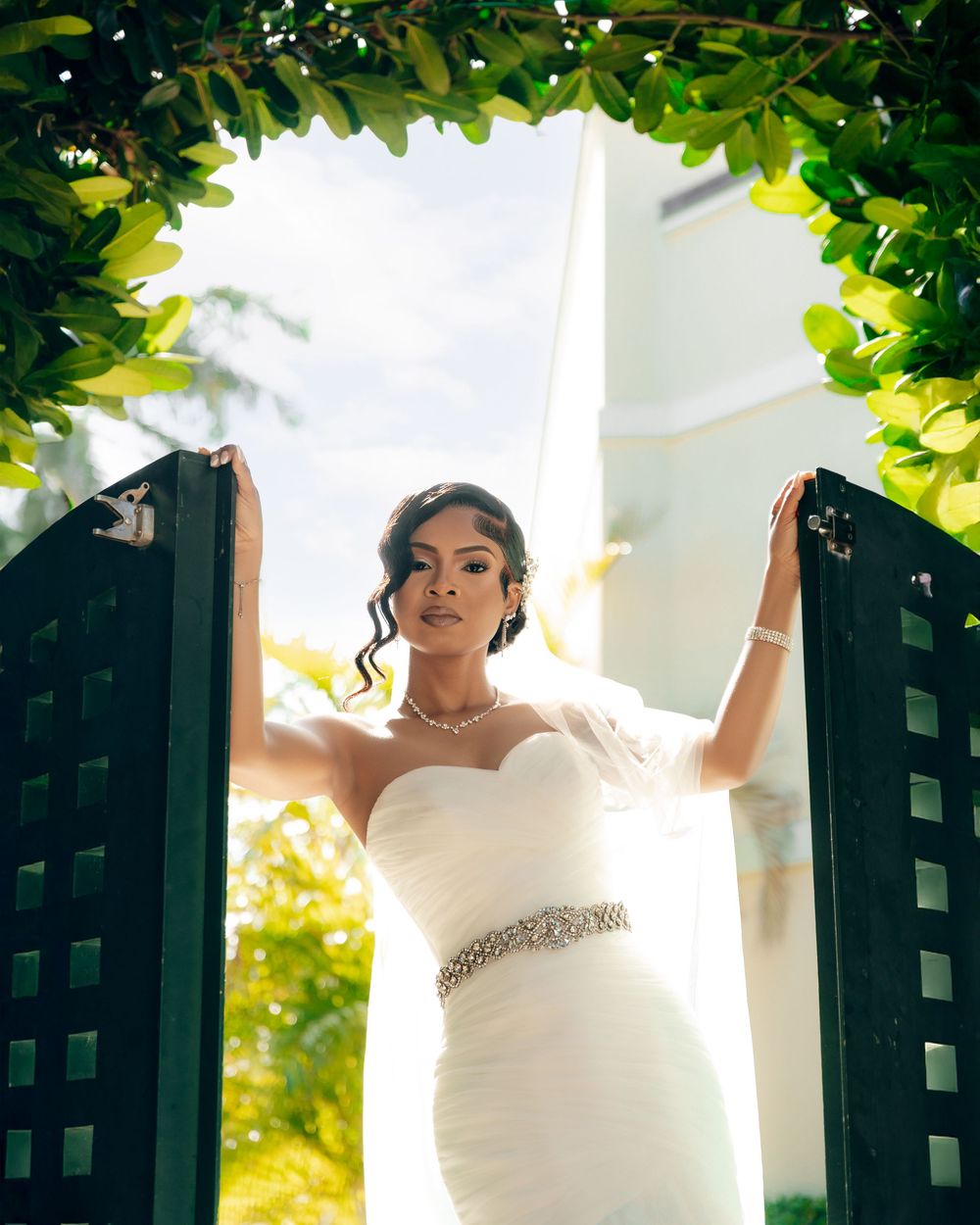 A bride in an elegant white fitted dress stands gracefully between garden gates framed by lush green foliage.