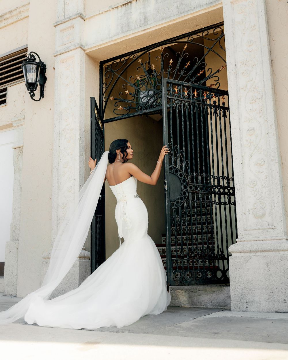 Elegant bride in white mermaid gown and flowing veil stands at ornate wrought iron gate entrance against cream building.