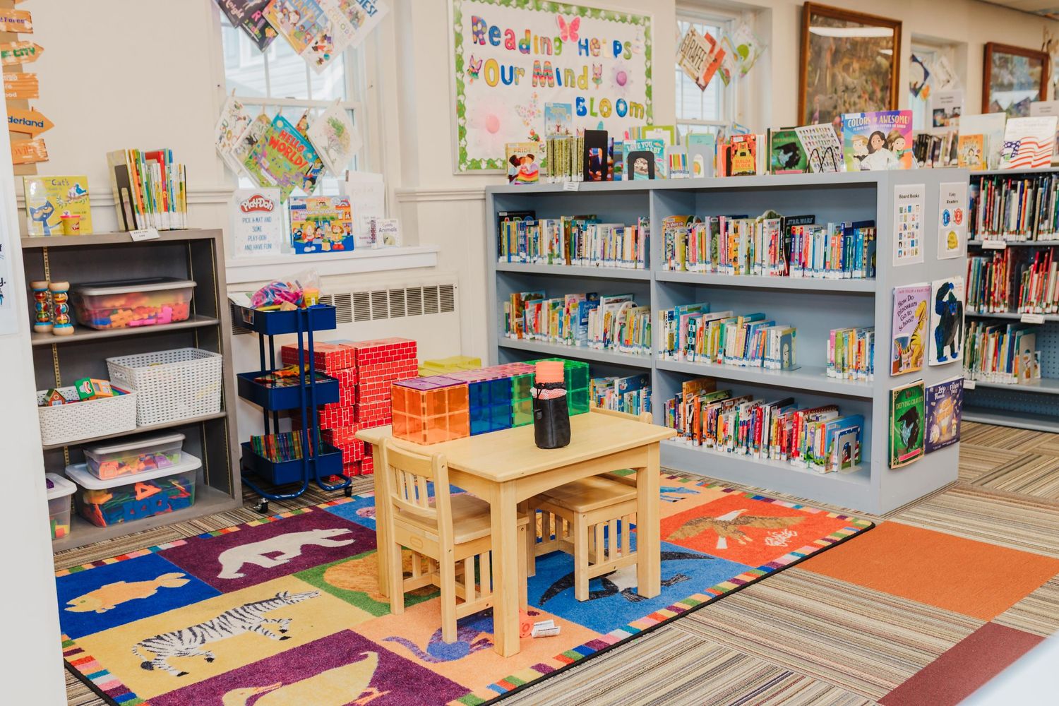 Colorful kids section library interior with bookshelves, reading area, activities, small wooden table and chairs on patterned carpet.