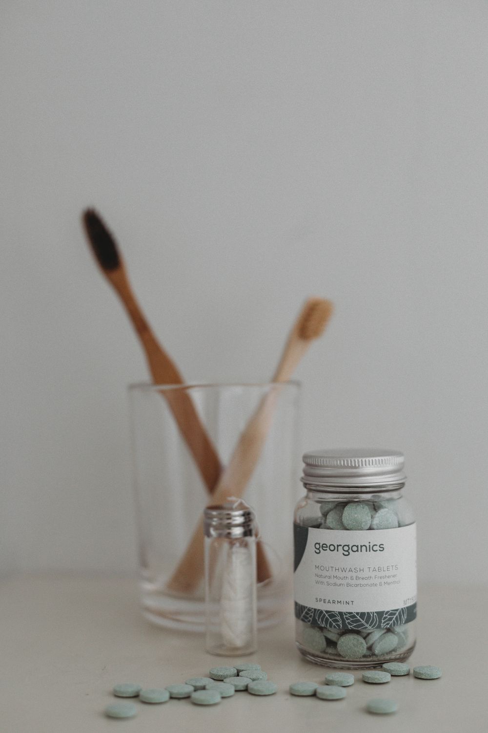 Glass jar of blue georganics dental tablets with bamboo toothbrushes in a glass on neutral background.