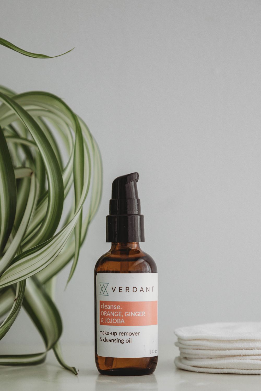 Amber glass bottle of Verdant cleansing oil displayed with cotton pads and green air plant on minimal white surface.