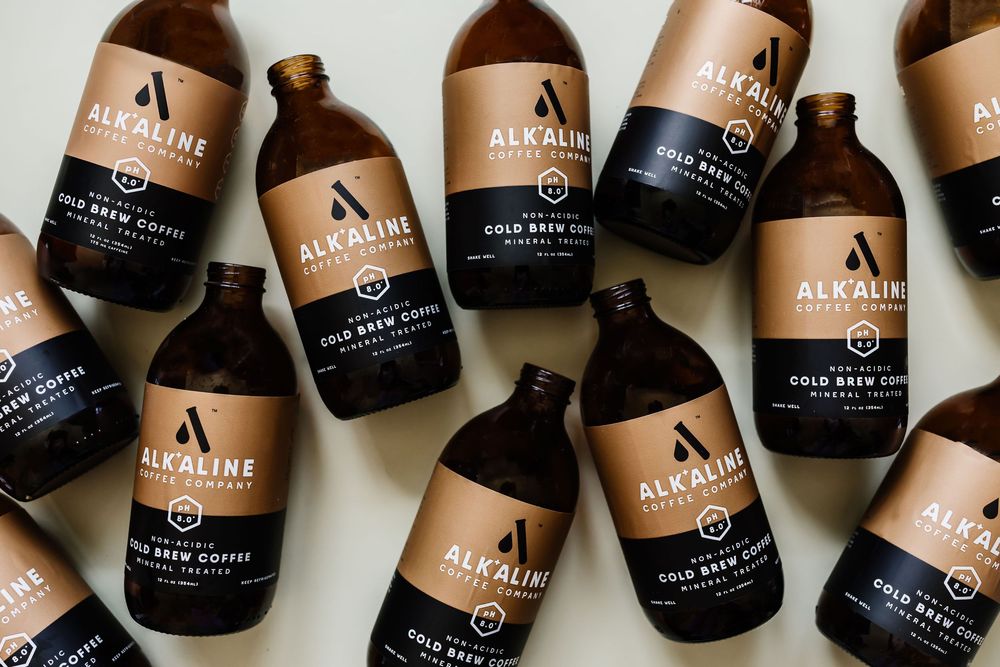 Multiple glass bottles of Alkeme brand cold brew coffee arranged in an artful pattern on dark background.