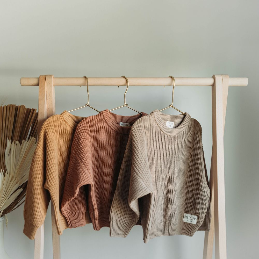 Three neutral-toned chunky knit sweaters hanging on wooden hangers displayed on a minimalist clothing rack.