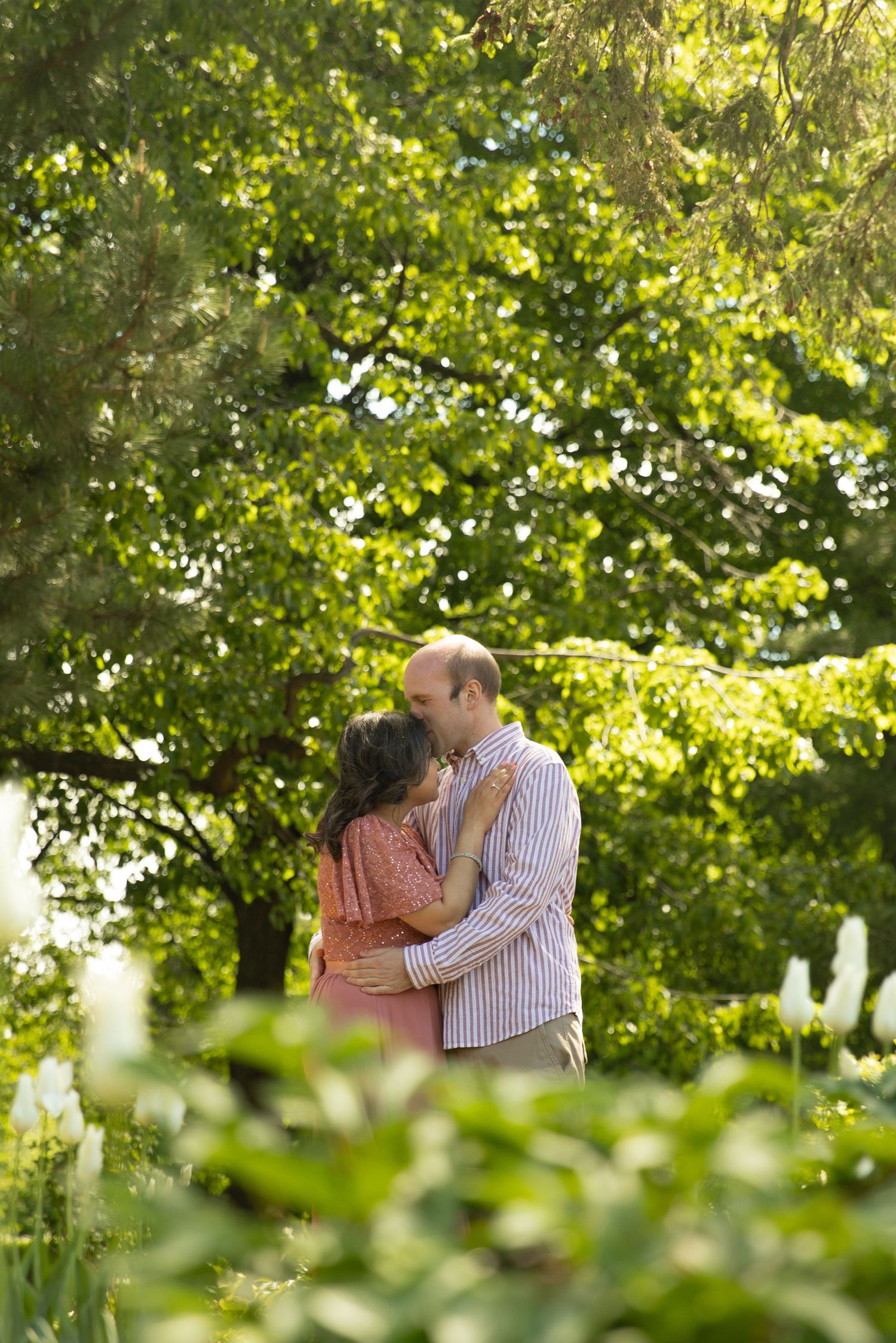 A couple shares a tender moment in a sunlit garden surrounded by lush green foliage and trees.
