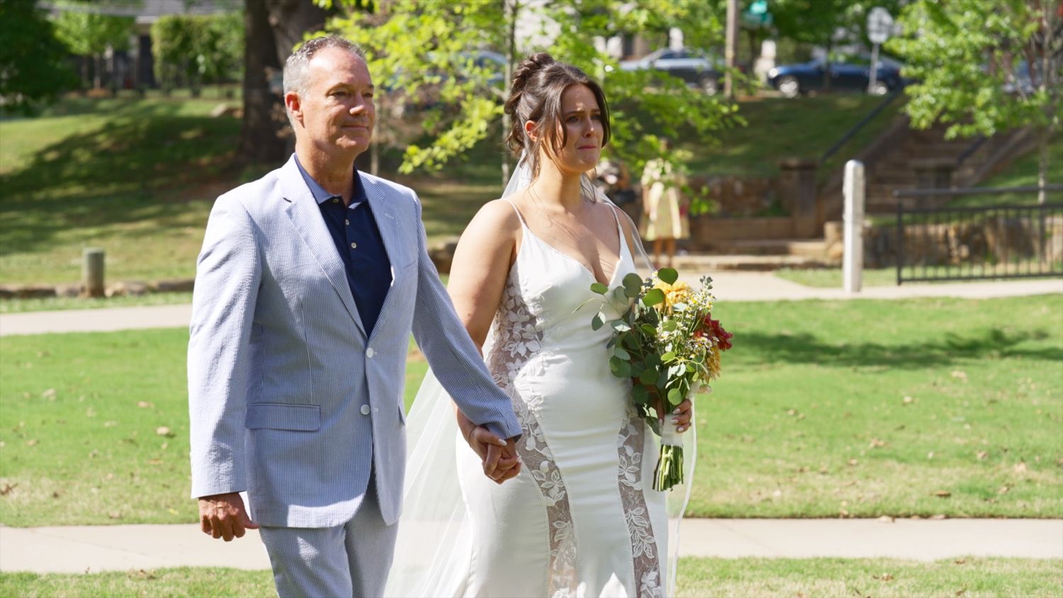A couple walks together at an outdoor wedding ceremony, with the bride wearing a fitted white dress and holding a bouquet.