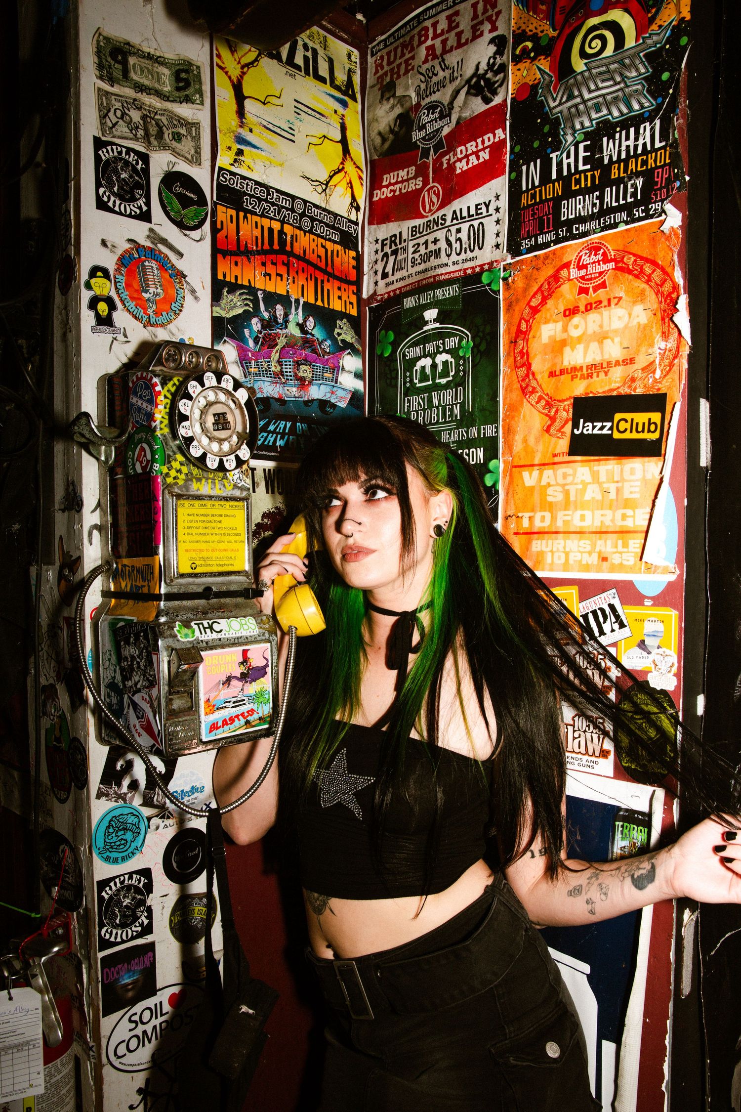Person in black outfit poses against wall covered in colorful concert posters and music flyers while holding yellow phone.