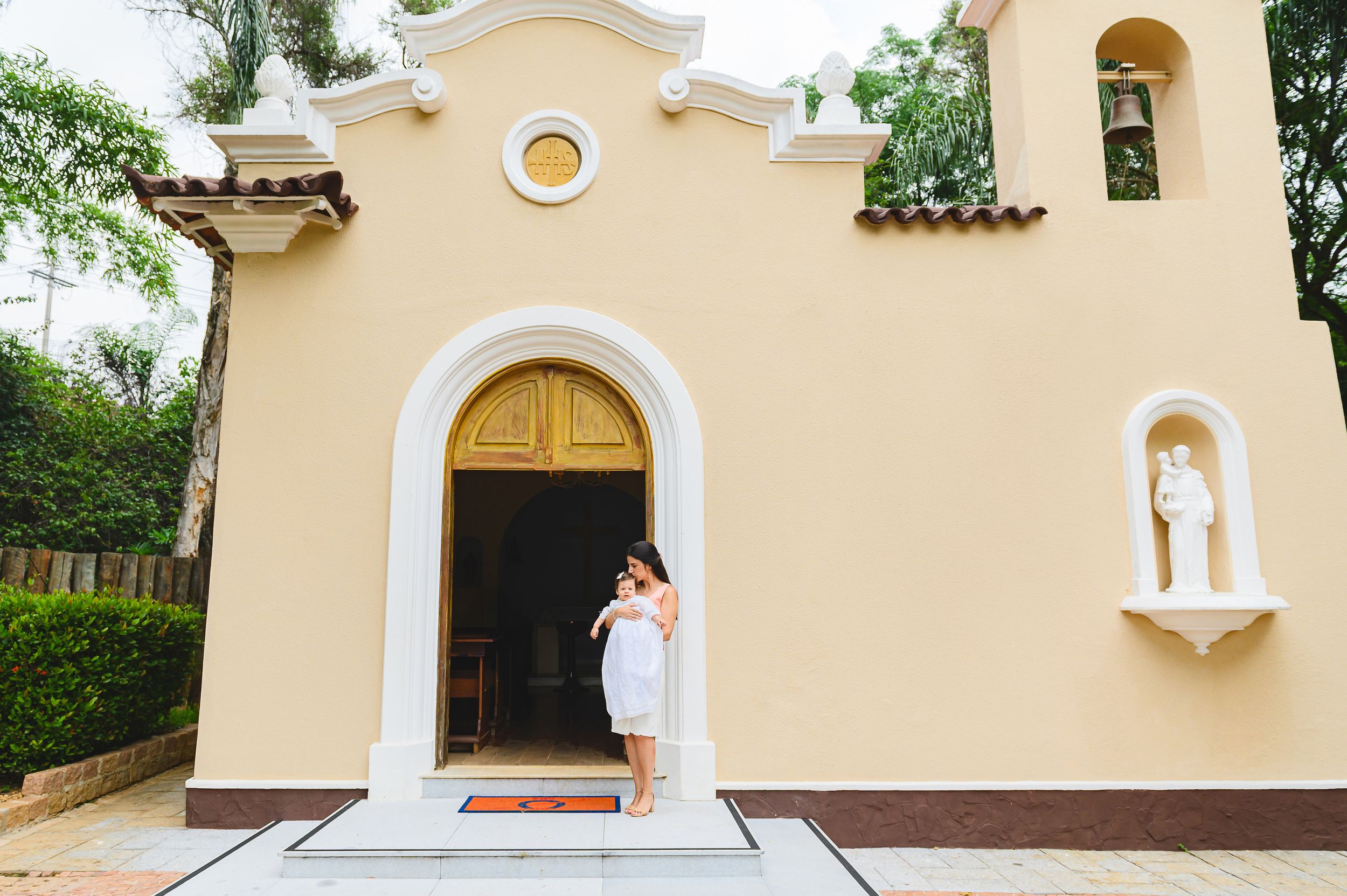 Small cream-colored chapel with arched doorway and white trim features classic Spanish mission architecture and lush greenery.