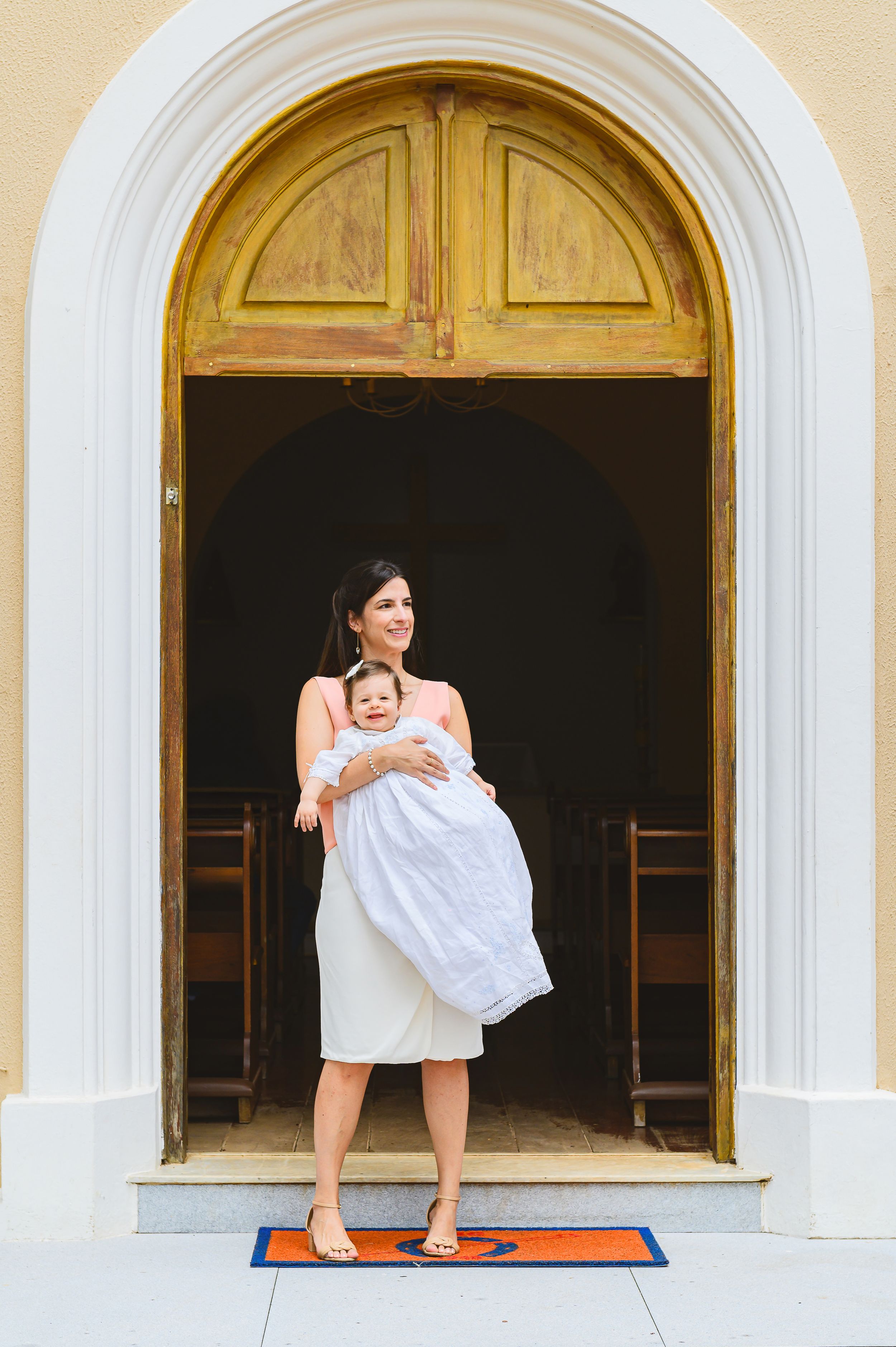 Someone stands in a white arched doorway of a church while holding a baby wrapped in white cloth.