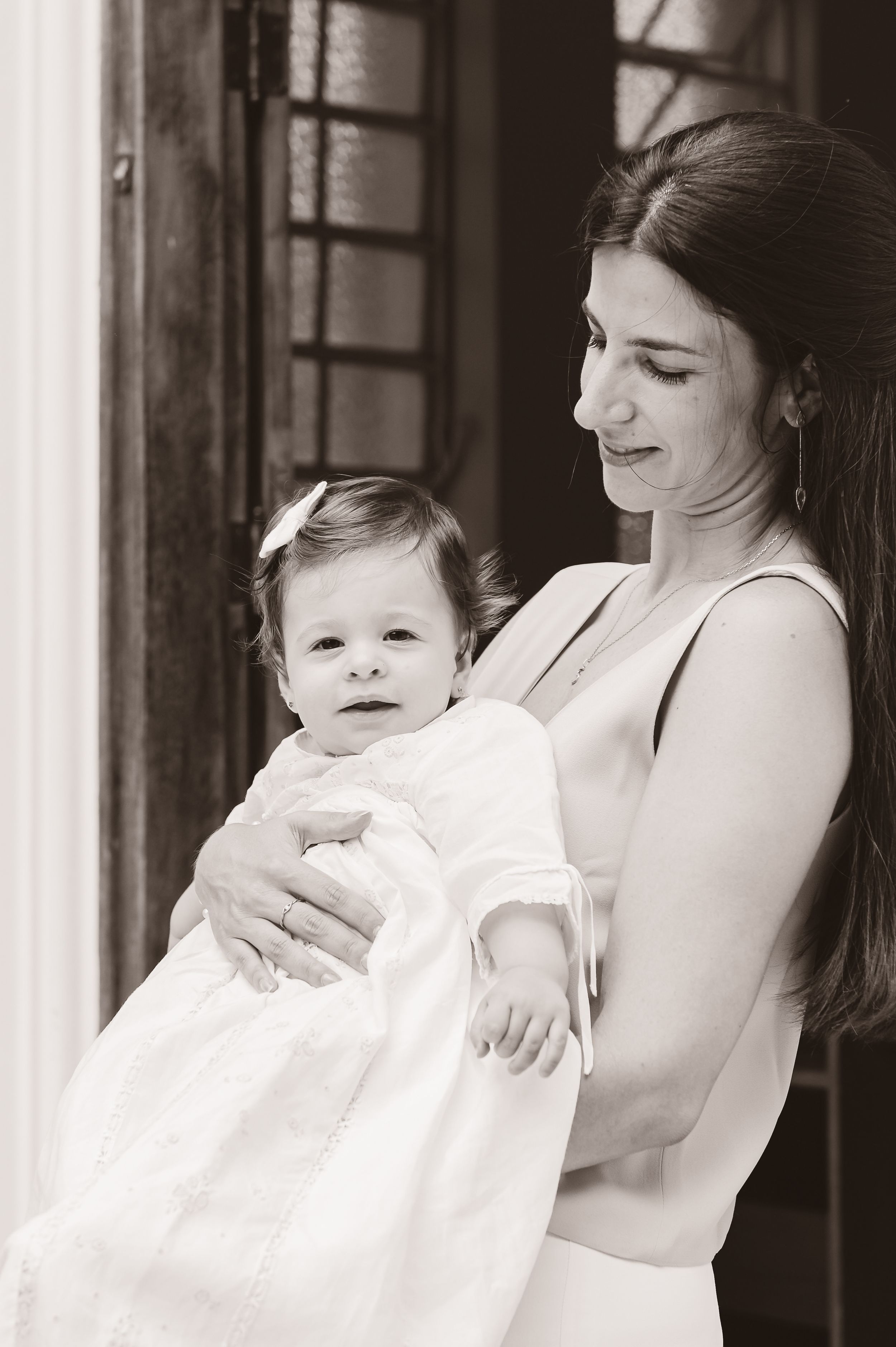 A black and white portrait of someone holding a baby in a white christening gown near a window.