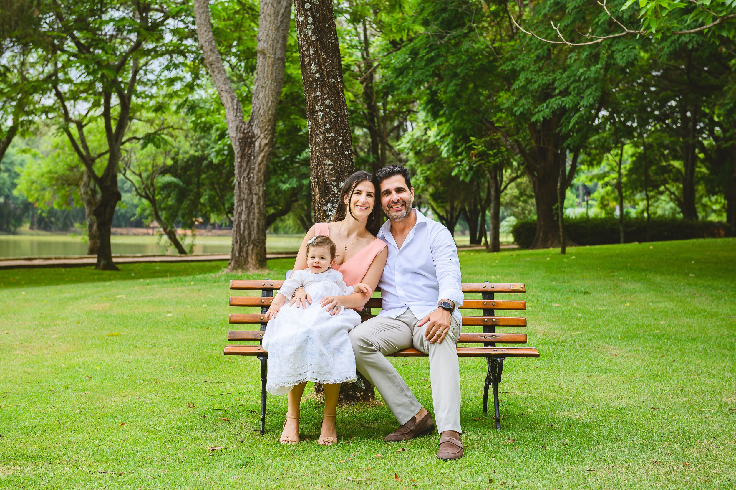Three people sitting together on a wooden park bench surrounded by lush green trees and grass in a peaceful setting.