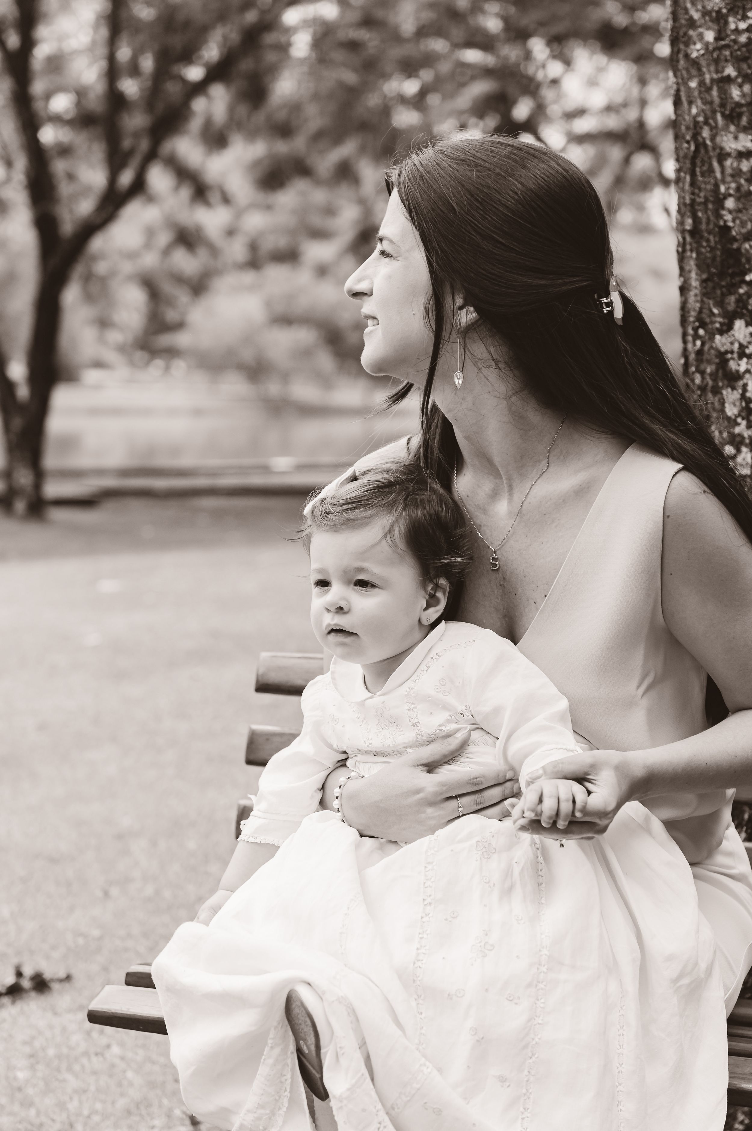 A tender black and white portrait of a caregiver and baby in a white dress sitting together on a park bench.