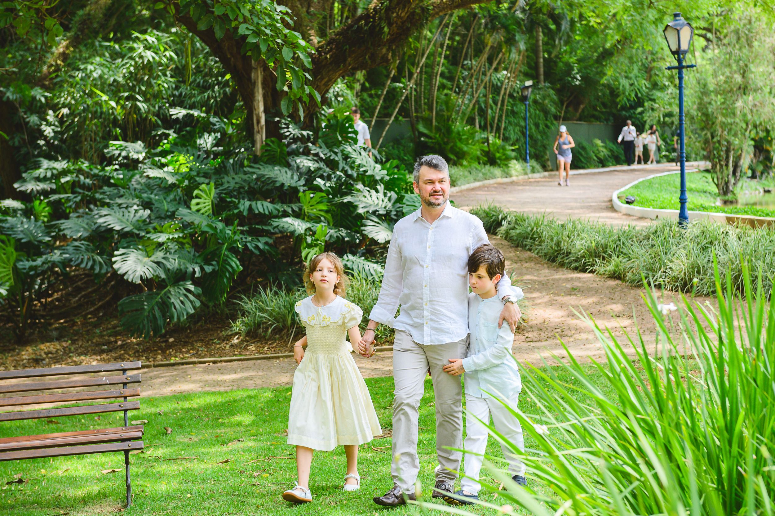 A person walks with two small children along a garden path surrounded by lush tropical plants and greenery.