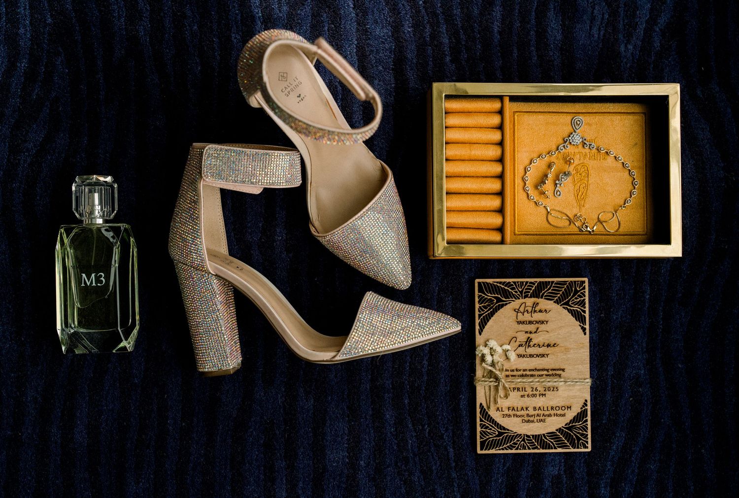 Wedding details flatlay featuring beige block heel sandals, perfume bottle, and gold invitation details on navy background.