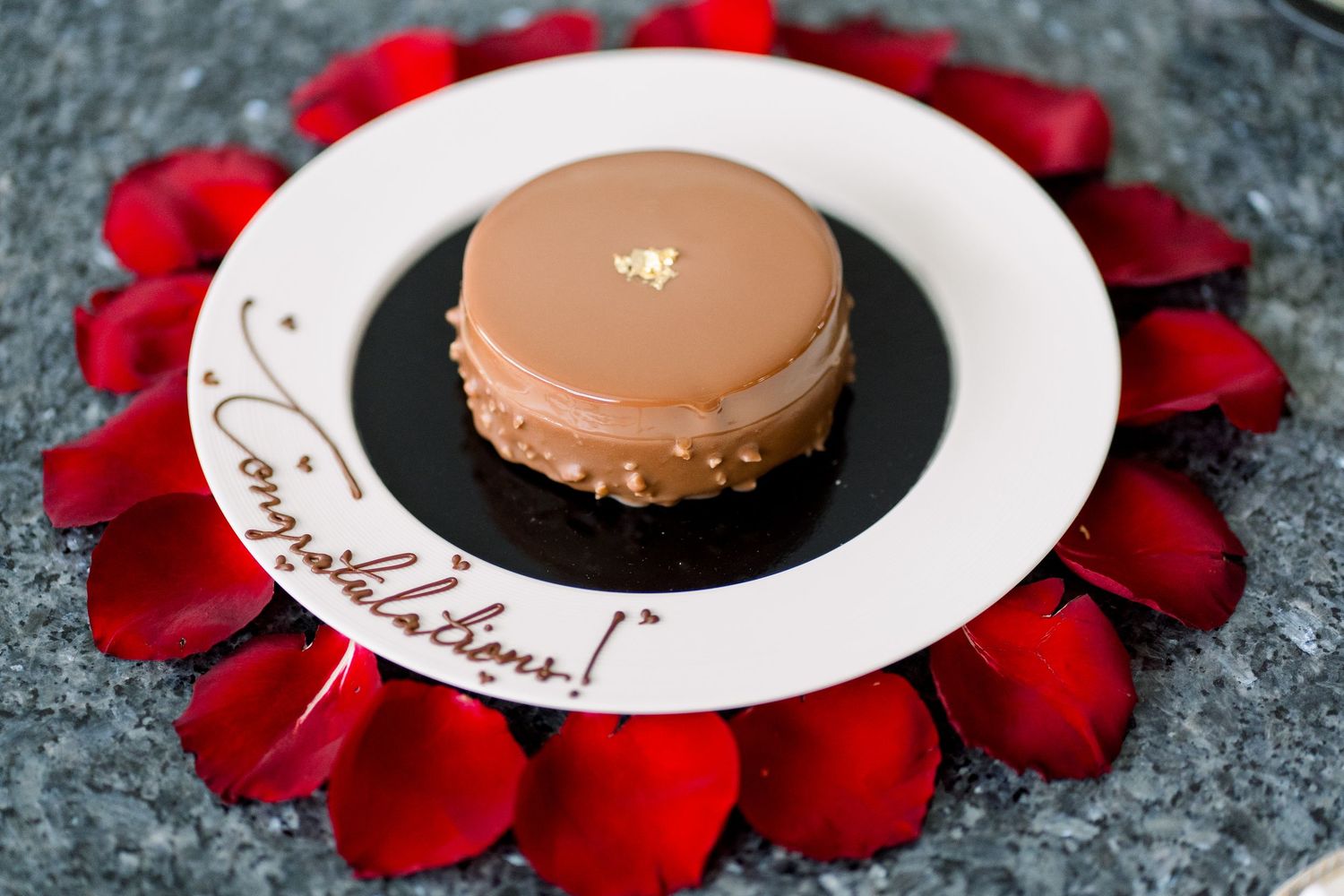 A round chocolate macaron sits on a black and white plate surrounded by red rose petals as decoration.
