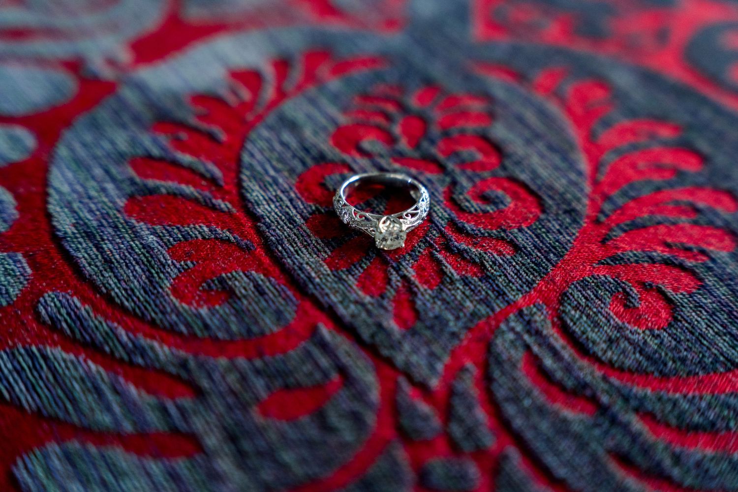 Diamond engagement ring placed on a red and black damask fabric with elegant floral patterns.