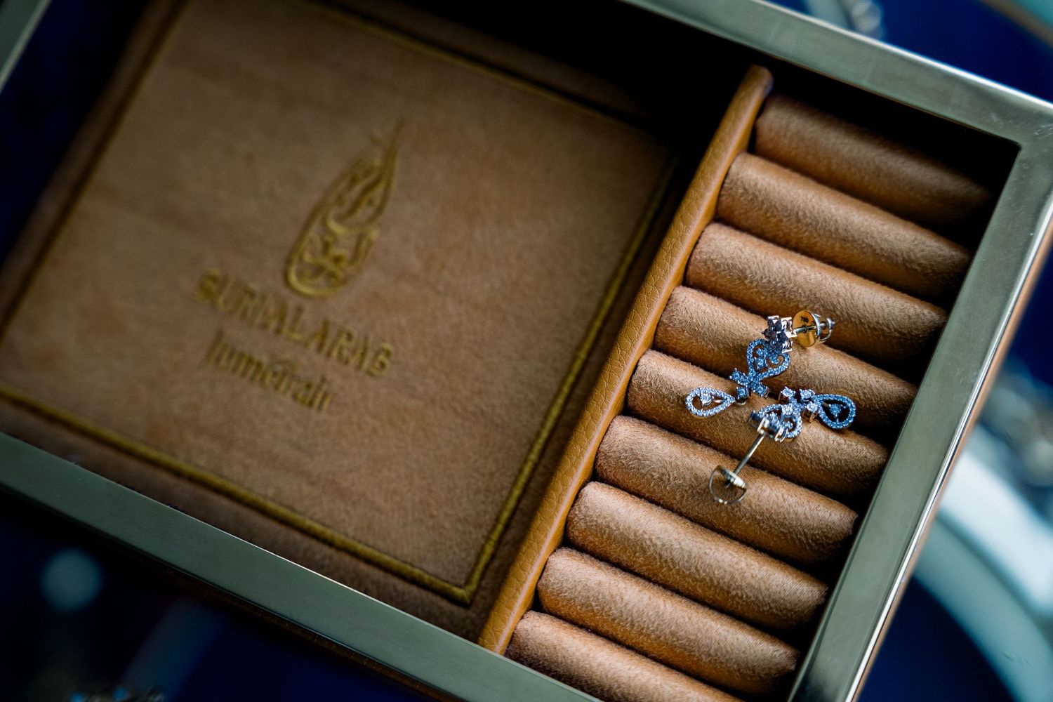 Elegant diamond ring sits among velvet ring holders inside a luxury wooden jewelry box with gold embossed logo.