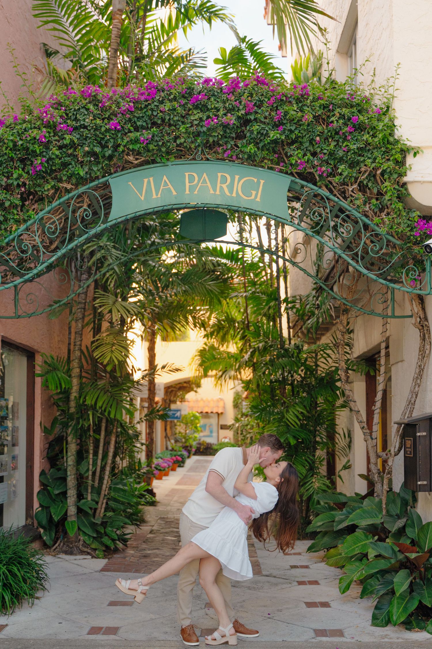 A romantic archway with 'Via Parigi' sign covered in blooming bougainvillea and tropical plants in an Italian courtyard at sunset.
