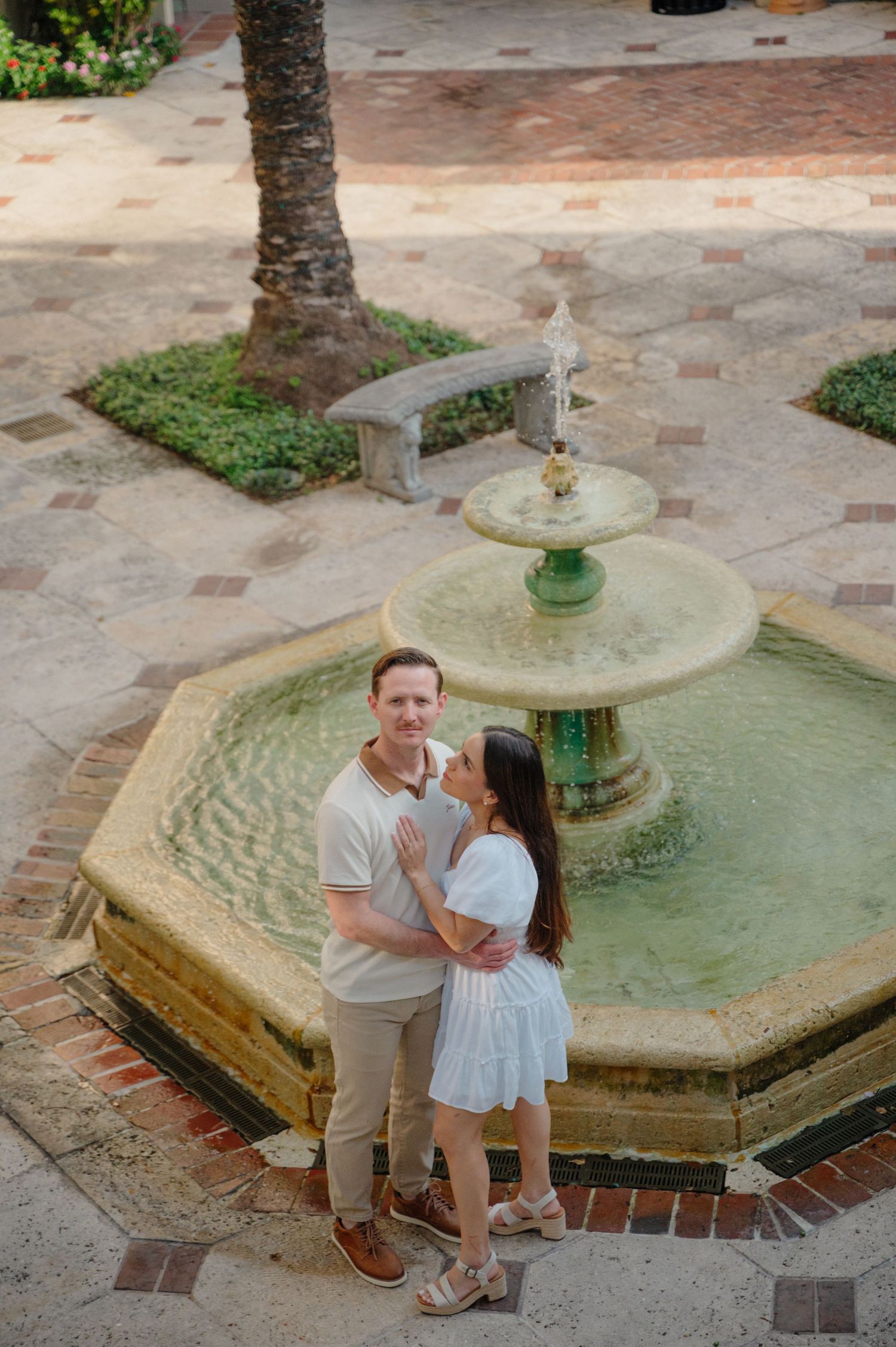 A couple stands together beside a decorative tiered fountain in a brick-paved courtyard with palm trees.