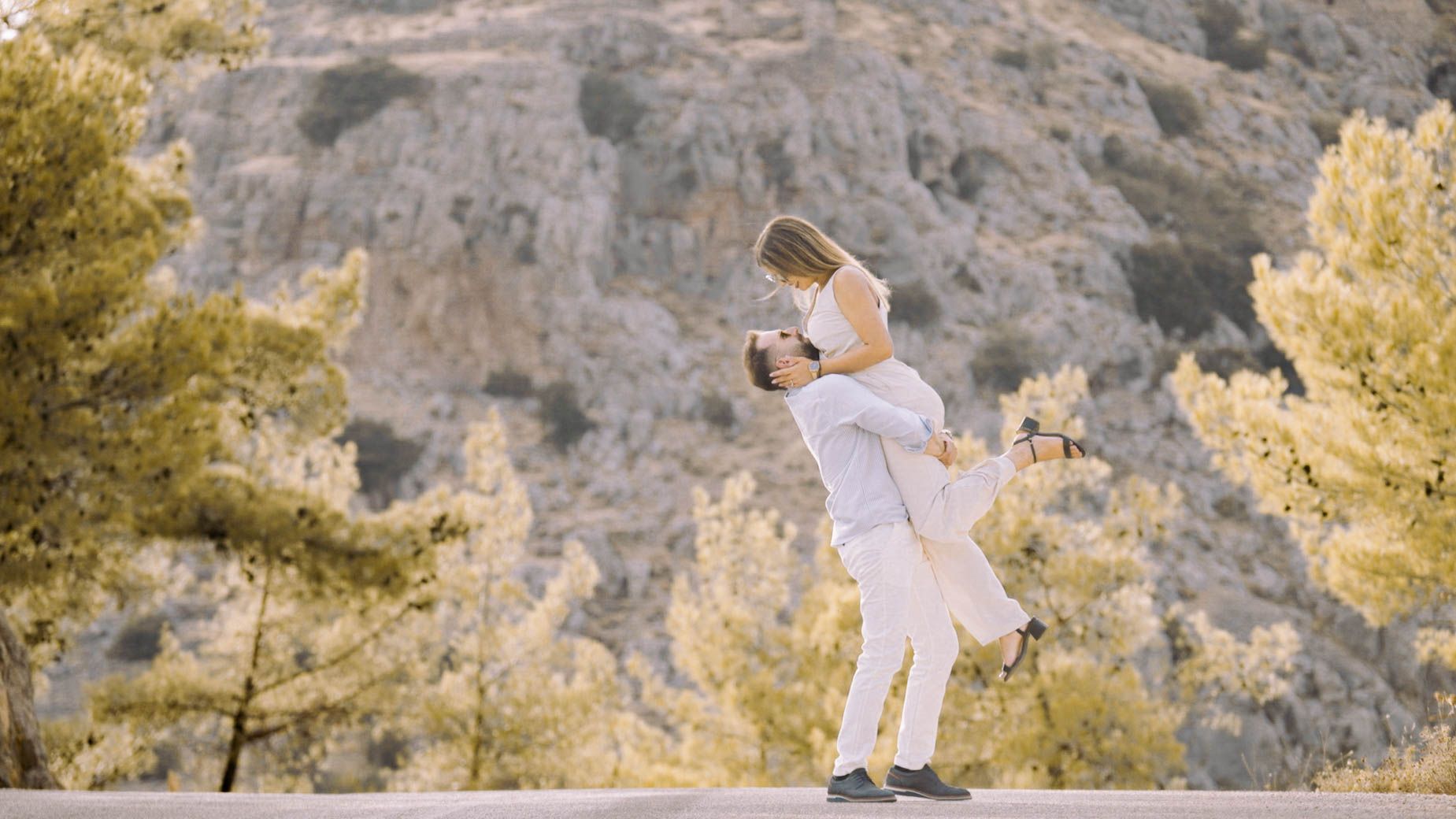 A romantic outdoor moment between a couple in white outfits surrounded by pine trees and rocky mountains.