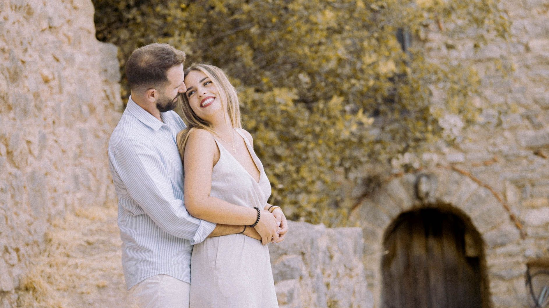 A romantic couple in light colored outfits embrace near a stone archway during a golden afternoon.