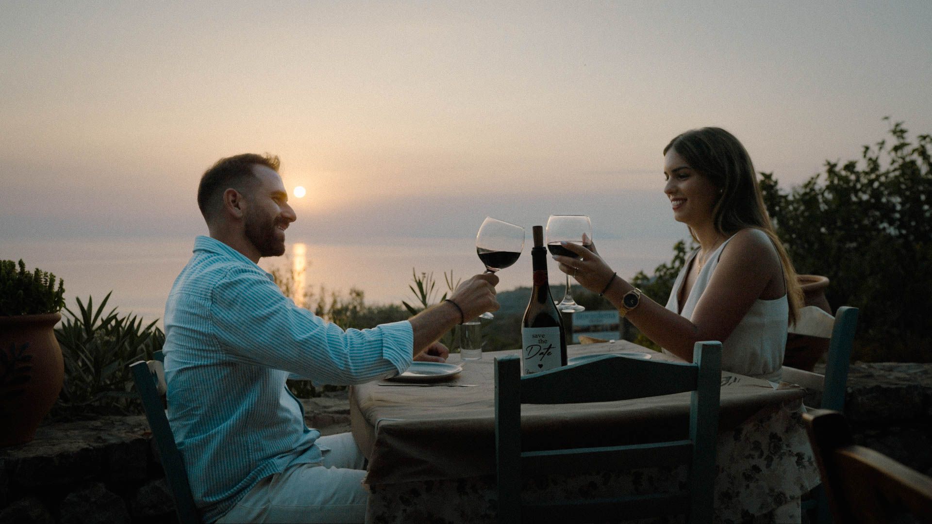 A couple toasts with wine glasses during a romantic outdoor dinner at sunset overlooking a scenic view.