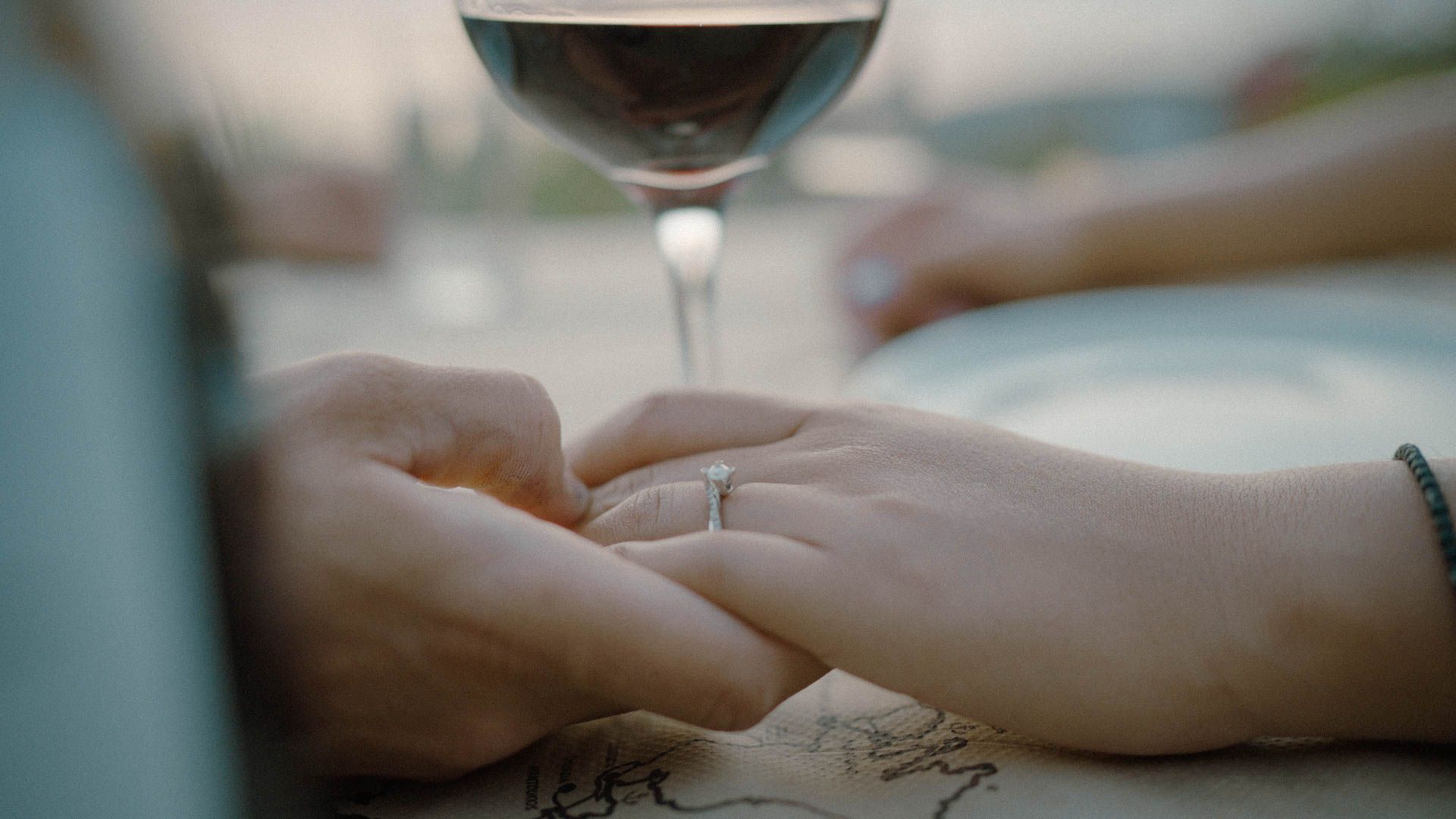 Two hands clasped together on a table with a glass of red wine in soft focus in the background.