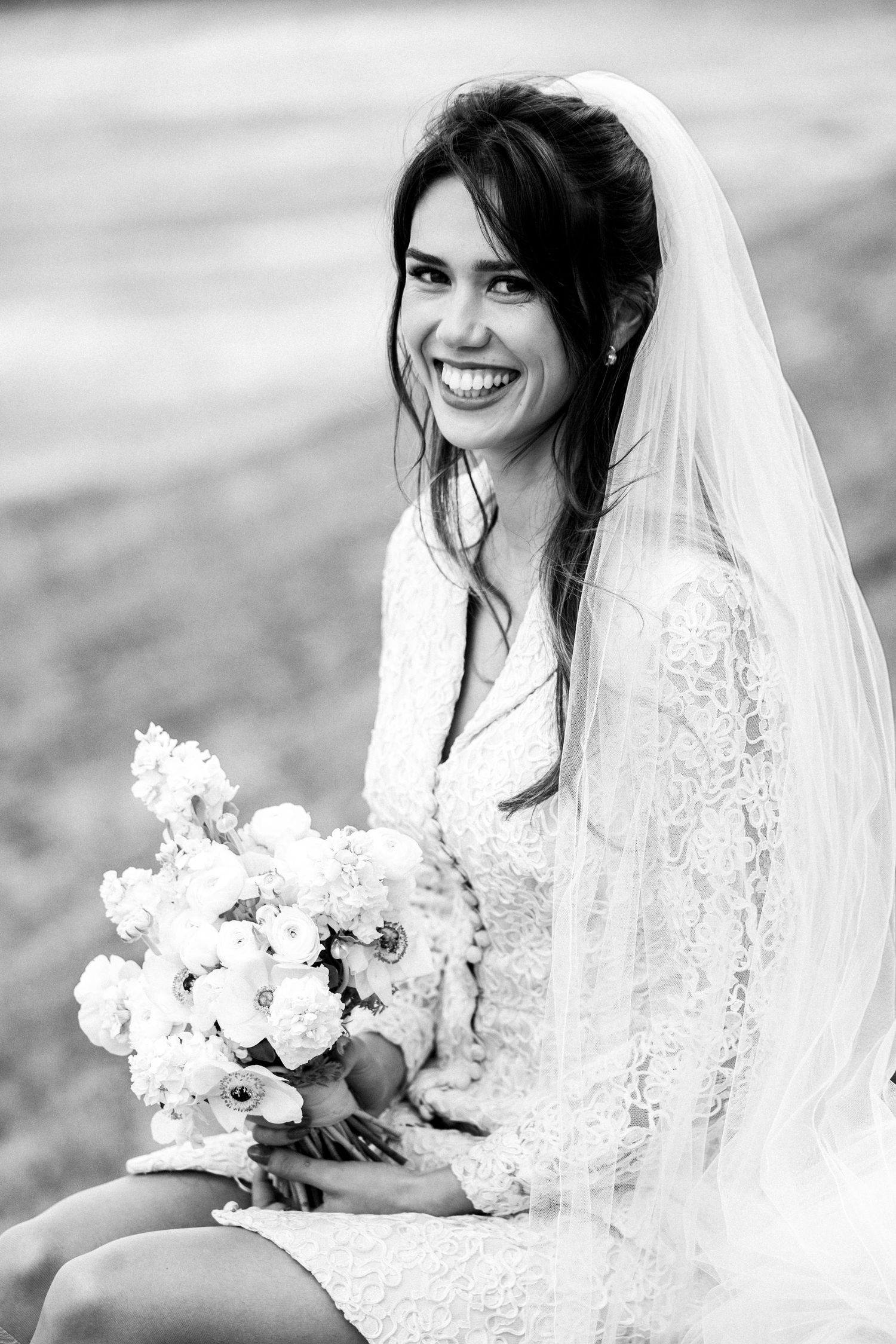 Black and white portrait of a bride wearing a lace wedding dress and veil while holding a white floral bouquet.