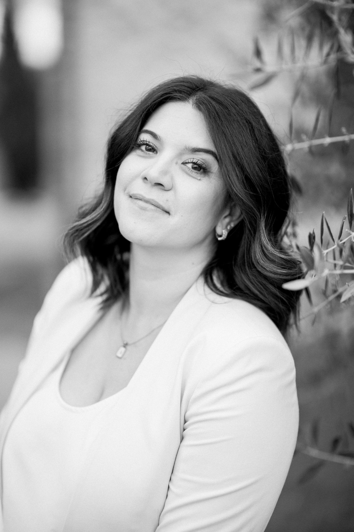 Black and white portrait of someone with long dark hair and a white top smiling softly against a blurred background.