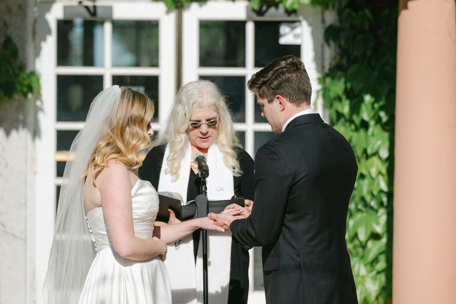 Three people stand together during an outdoor wedding ceremony in front of a white building with greenery.