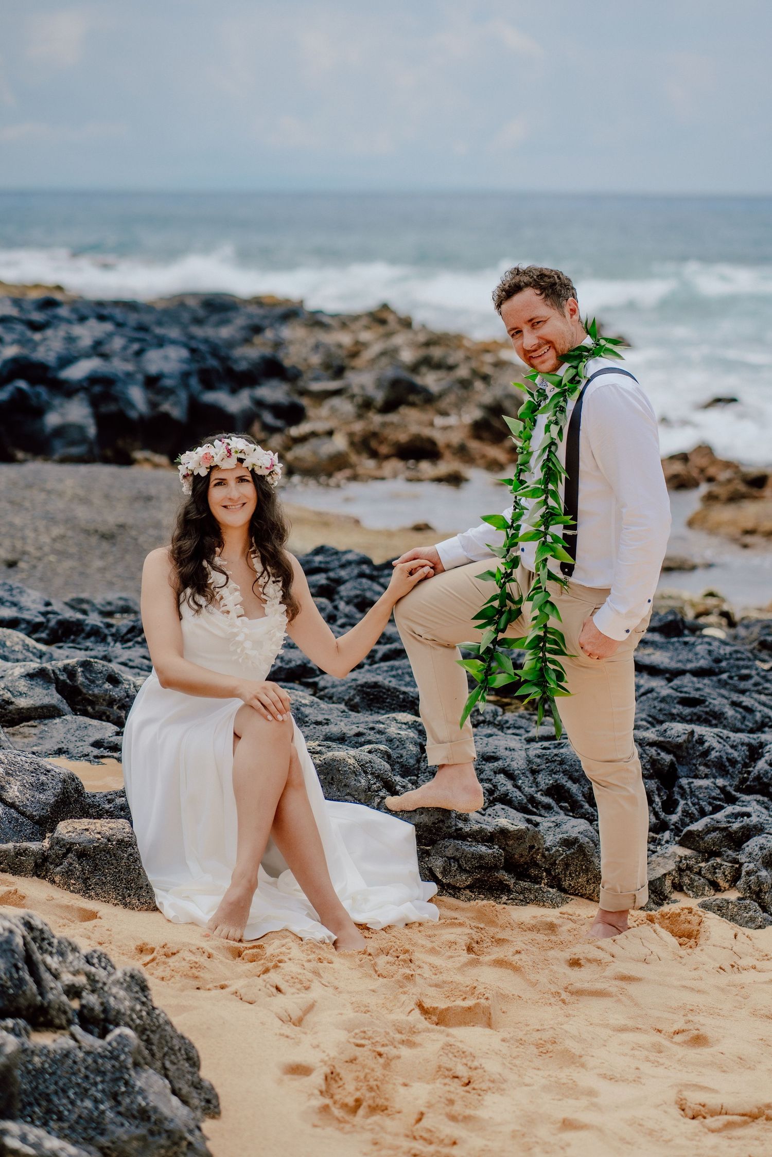 A couple poses on a rocky beach with waves in background, bride in white dress and groom wearing green leaf lei.