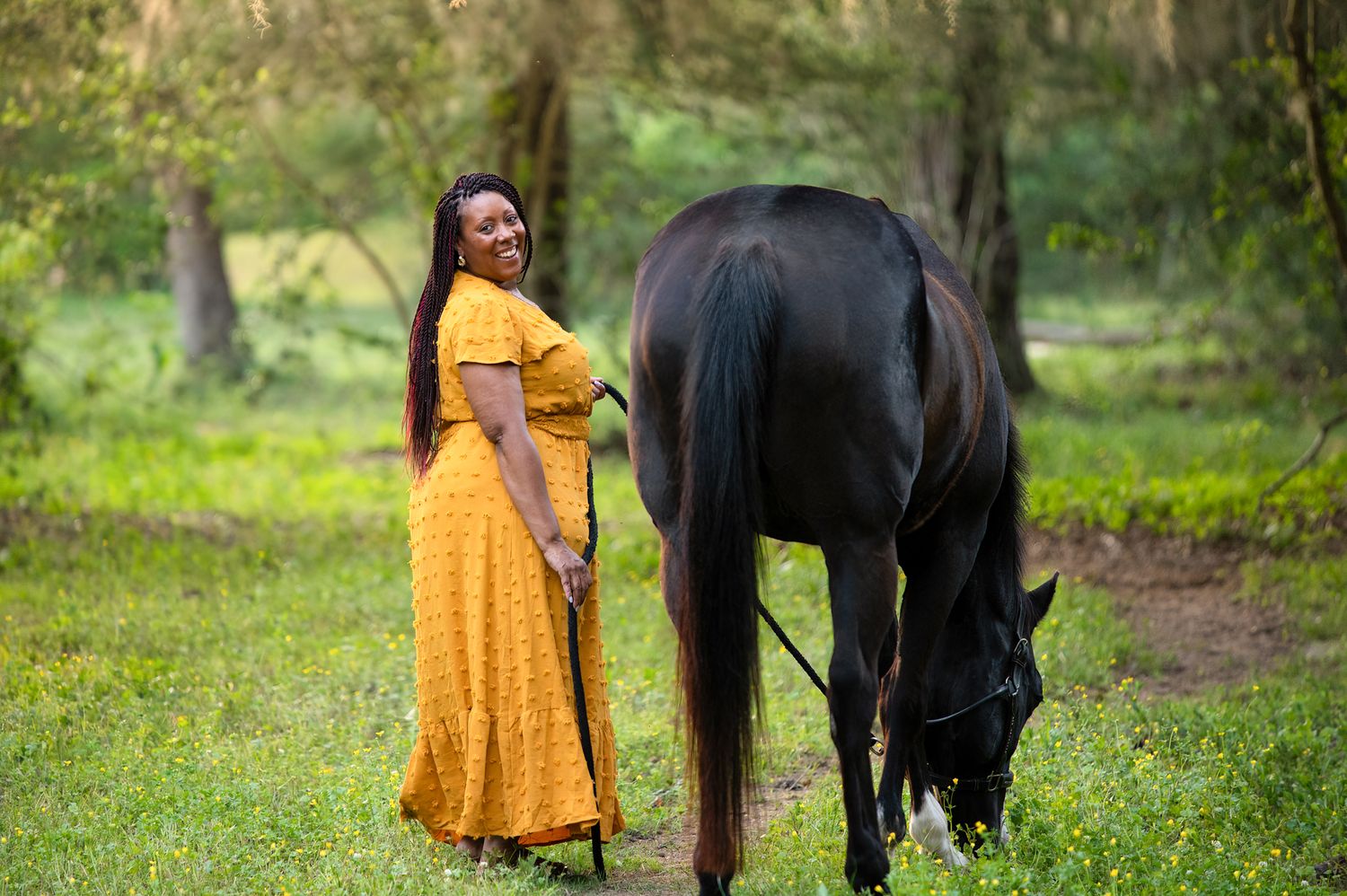 Woman wearing a yellow polka dot dress stands in a green forest meadow beside a black horse grazing.