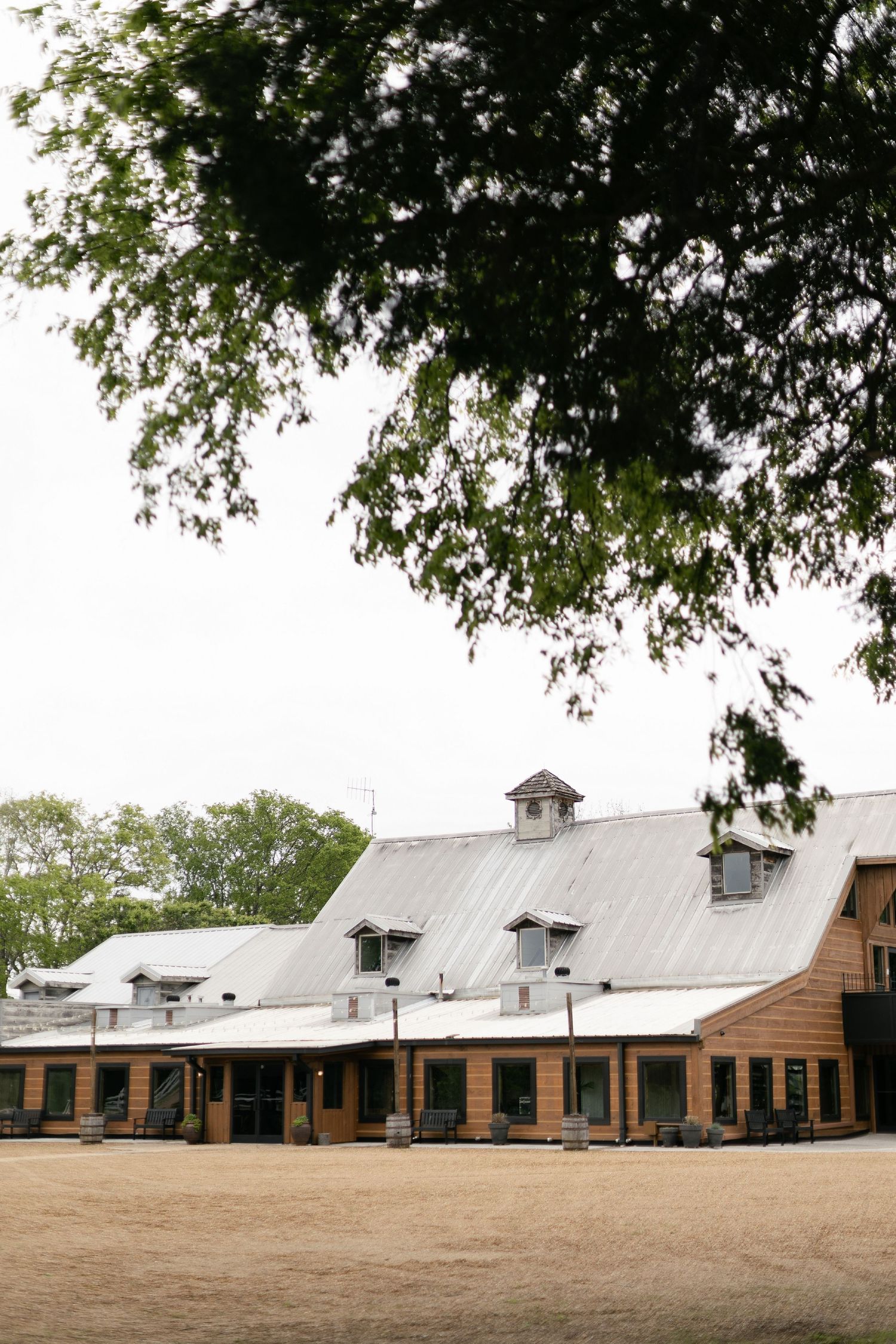 A rustic equestrian facility with a metal roof and wooden siding sits beneath overhanging tree branches on a dirt paddock.