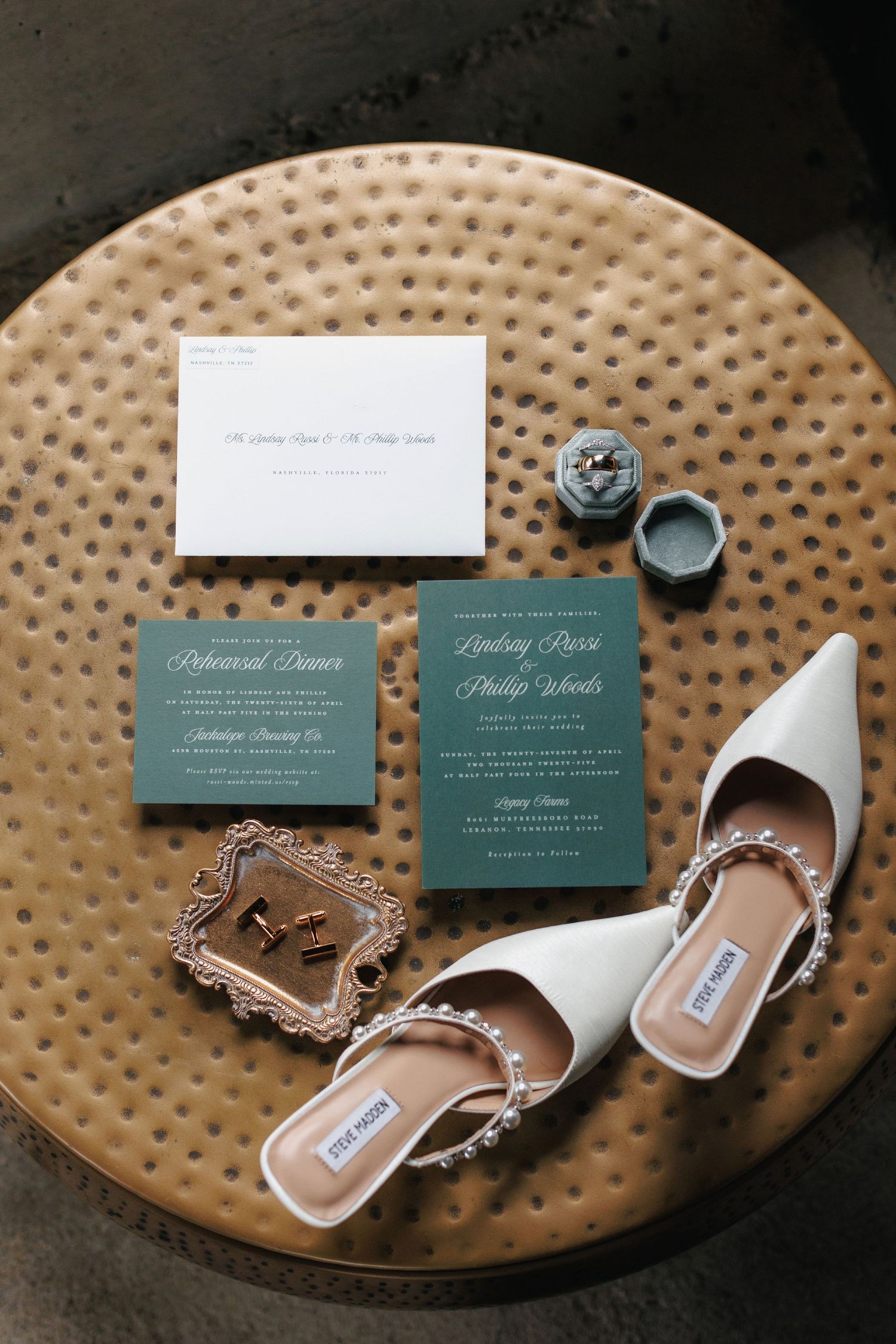 Wedding invitation cards and elegant white bridal shoes arranged on a textured round metal surface.