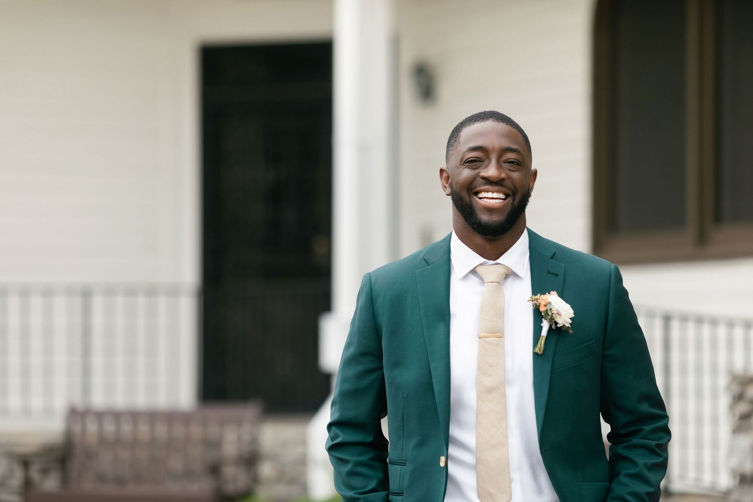 Groom wearing emerald green suit jacket and white boutonniere smiles joyfully outdoors.