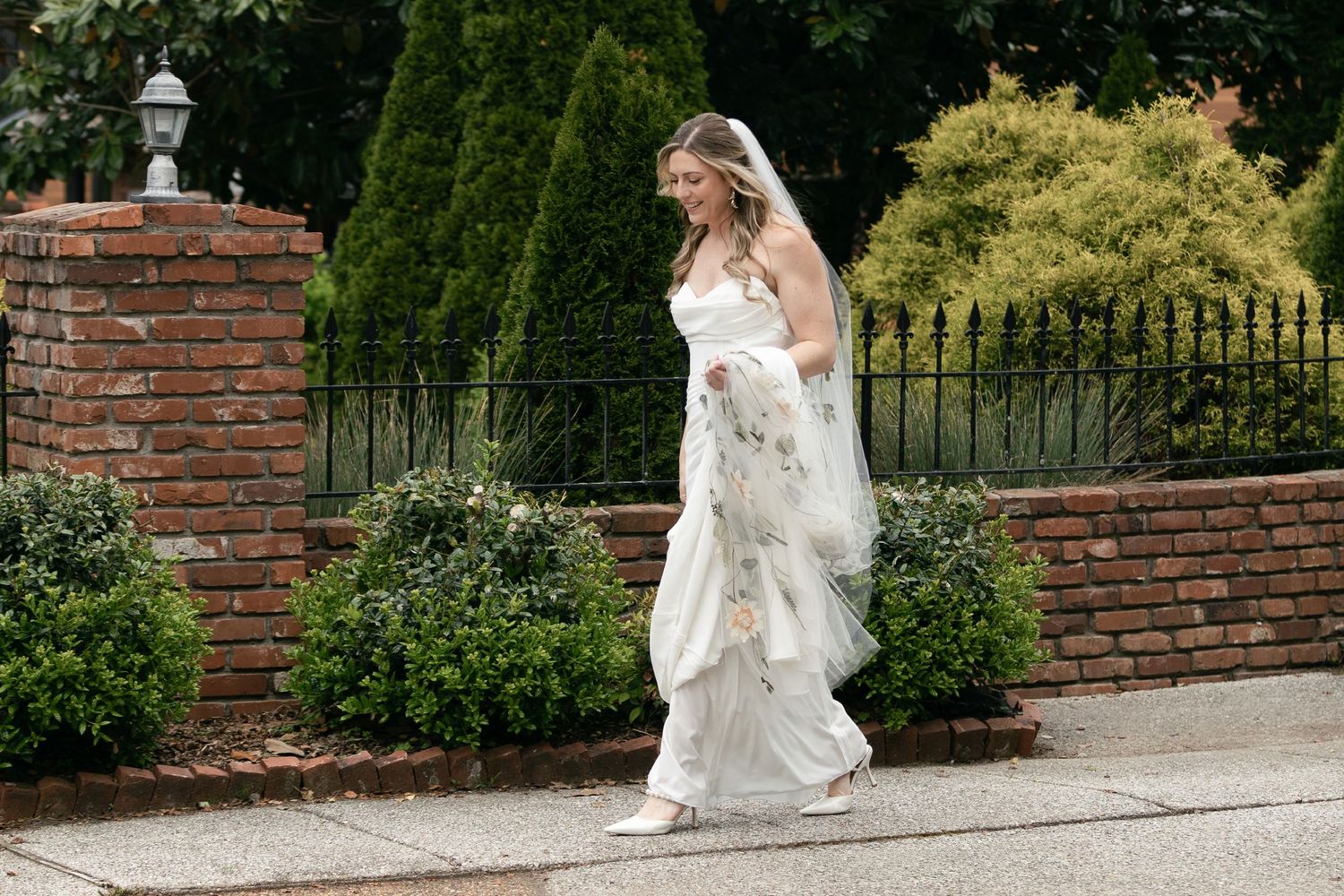 A bride in a flowing white wedding dress walks beside a brick wall and manicured garden on her special day.