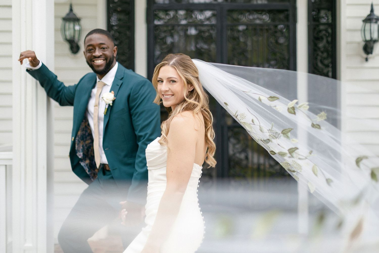A joyful couple on their wedding day with the bride in white and her veil flowing in the wind outside a white building.
