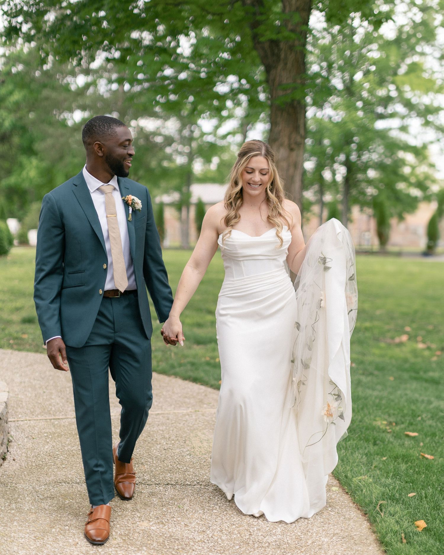 A joyful couple walking hand in hand on a garden path, the bride in a flowing white gown and groom in a dark suit.