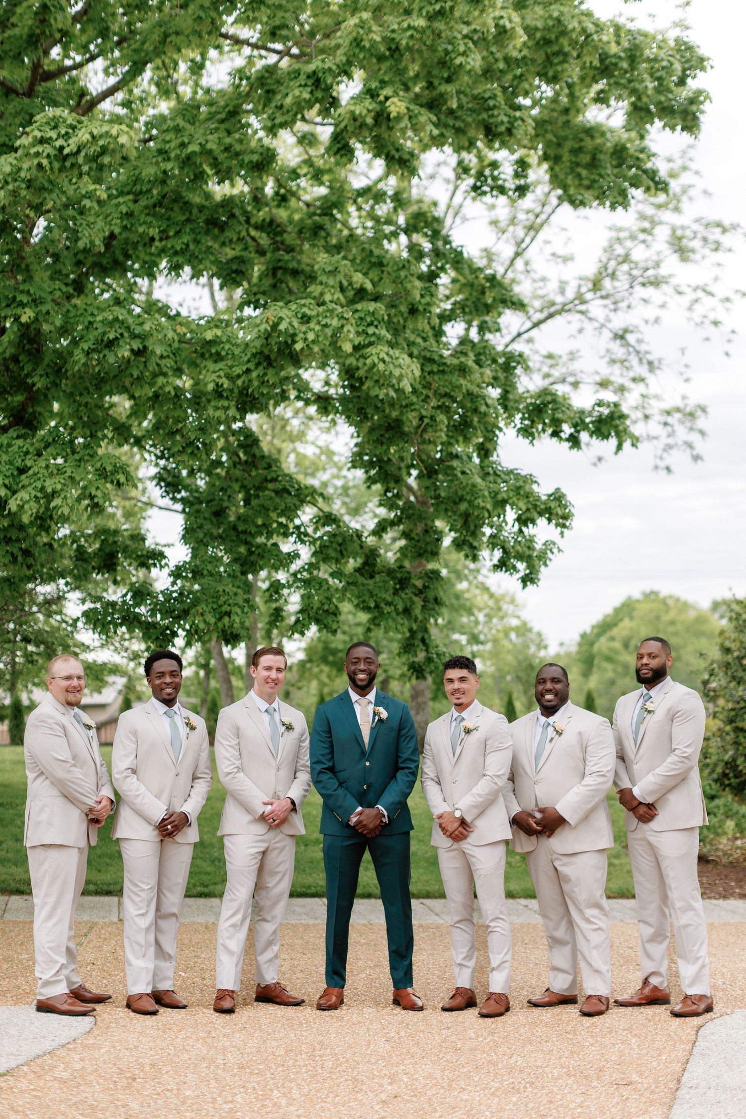 Groomsmen in light beige suits stand together with one person in middle wearing teal suit under leafy green tree.